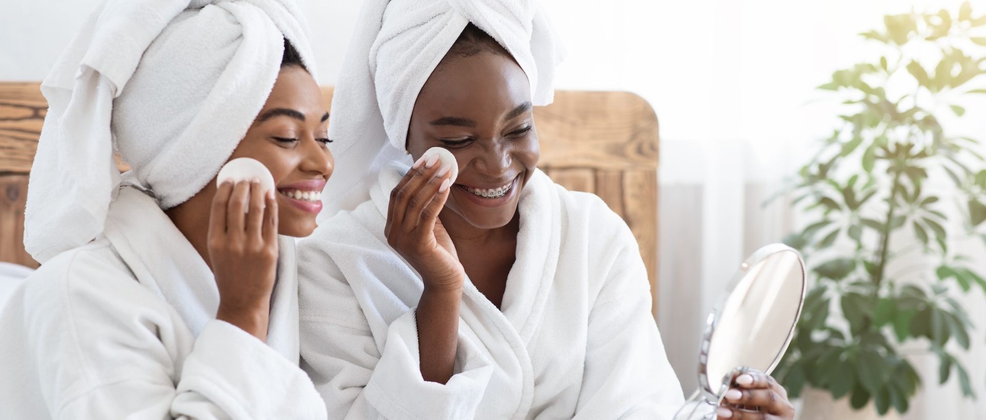 Smiling black female friends testing beauty products at home, using cotton pads, looking at mirror, bedroom interior, sun flare. African american ladies in bathrobes cleansing faces