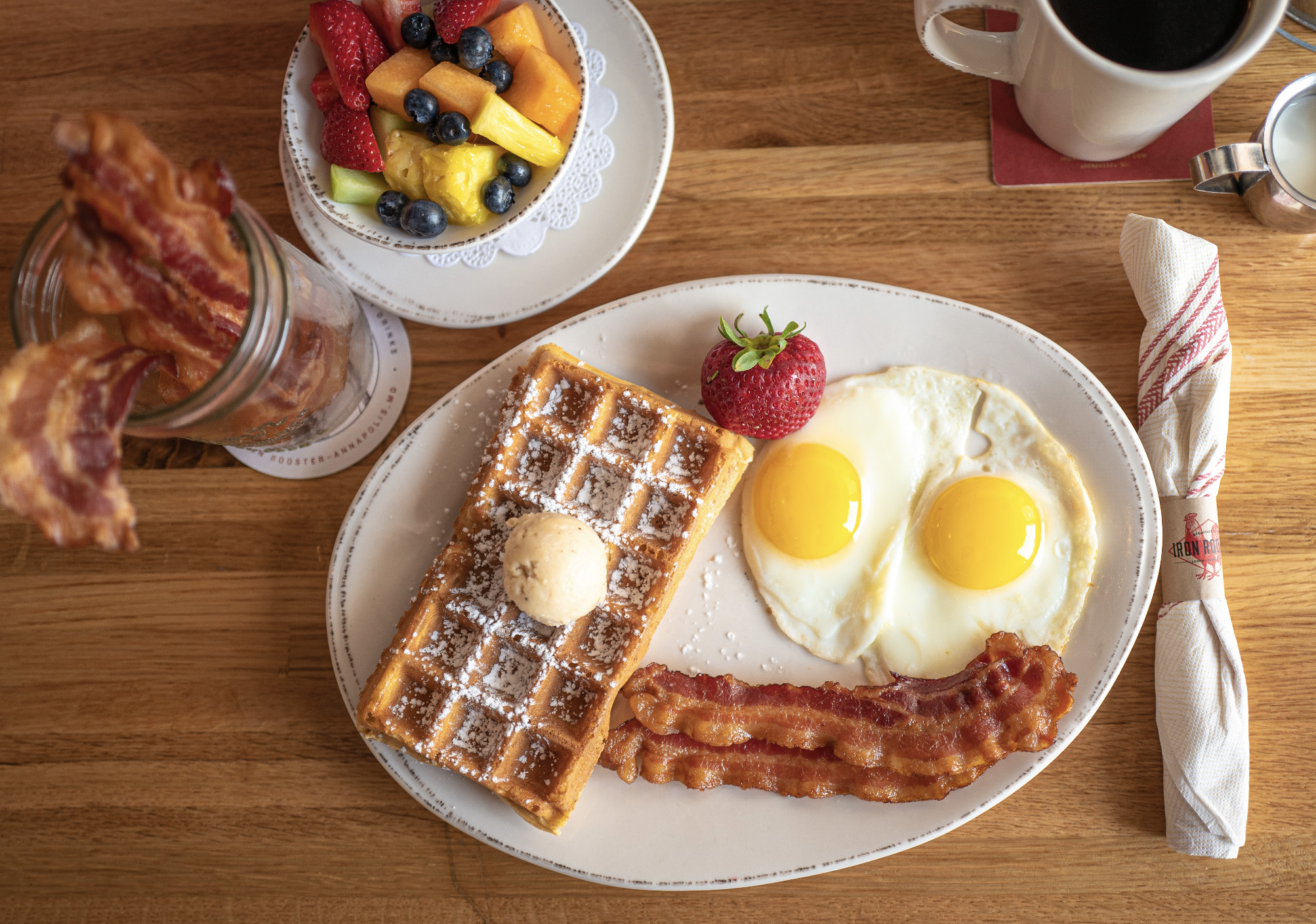 Full table breakfast at Iron Rooster including coffee, a bowl of fresh fruit, jar of Iron Rooster's famous candied bacon and a plate with Belgian waffles and two sunny side up eggs.